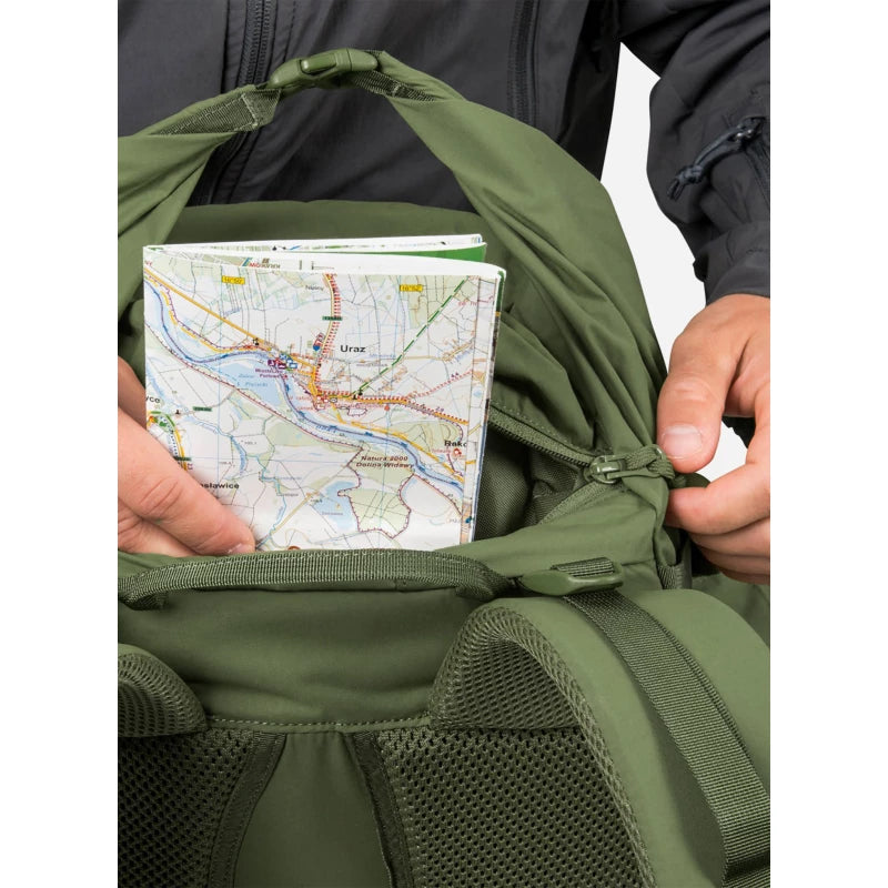 An olive green backpack shown close-up with a person's hands placing a paper map into the front pocket. The person is wearing a dark gray jacket, and the background is plain white.