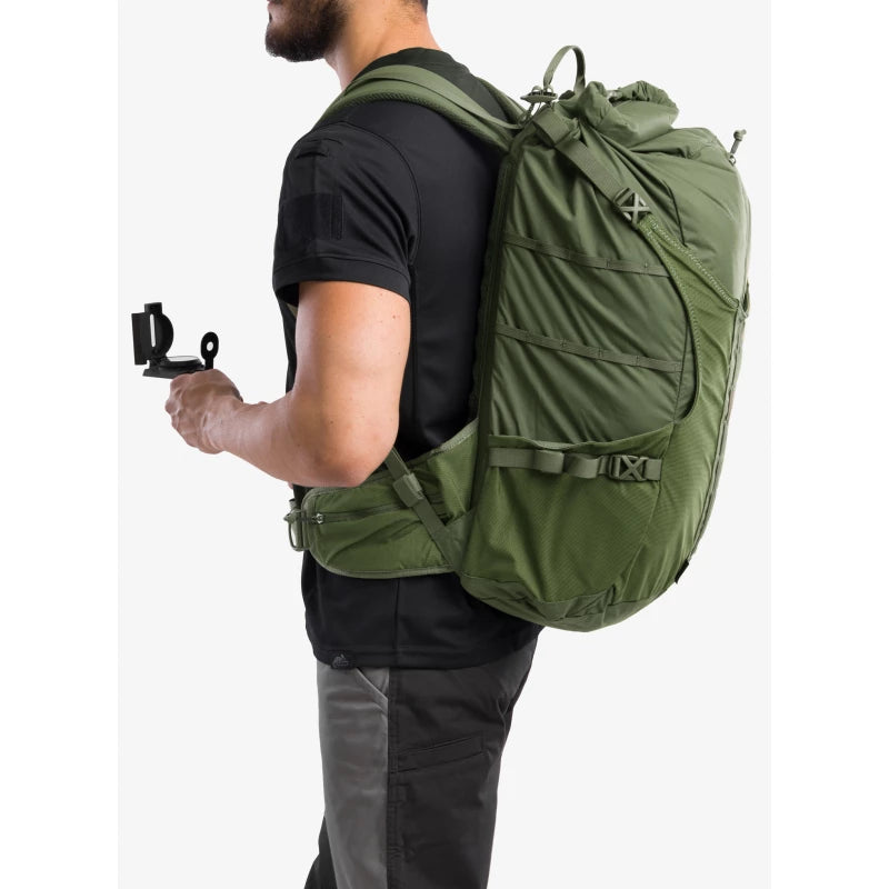 A man wearing a black t-shirt and dark pants is shown from the side/back wearing an olive green technical backpacking backpack. He is holding a black mounting bracket against a clean white background.