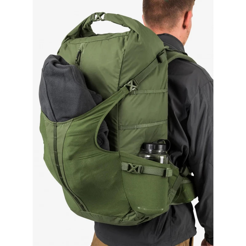 An olive green tactical backpack shown from the back on a model wearing a black shirt, with a gray fleece and a water bottle in the side pockets.