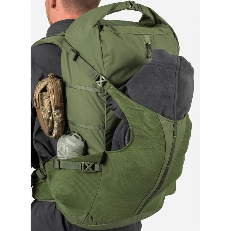 An olive green tactical backpack shown on a model from the back, carrying a dark gray fleece and a small camouflage pouch on the side.