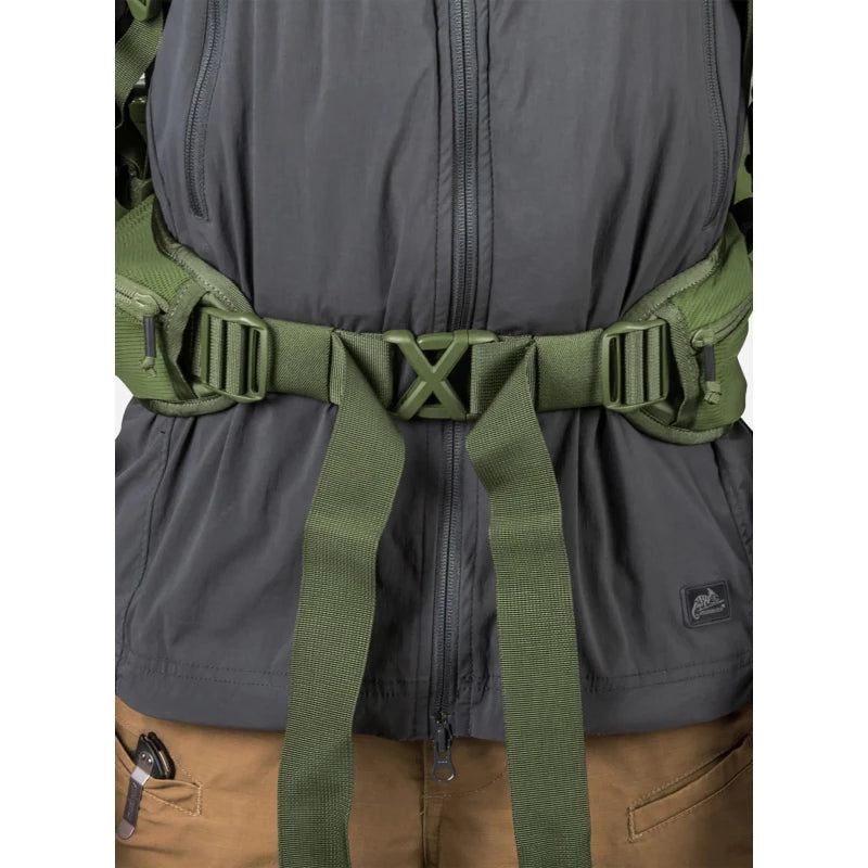 Close-up of a dark gray zip-front jacket with an olive green harness-style waist strap and buckle system worn across the torso, plus tan pants visible below.